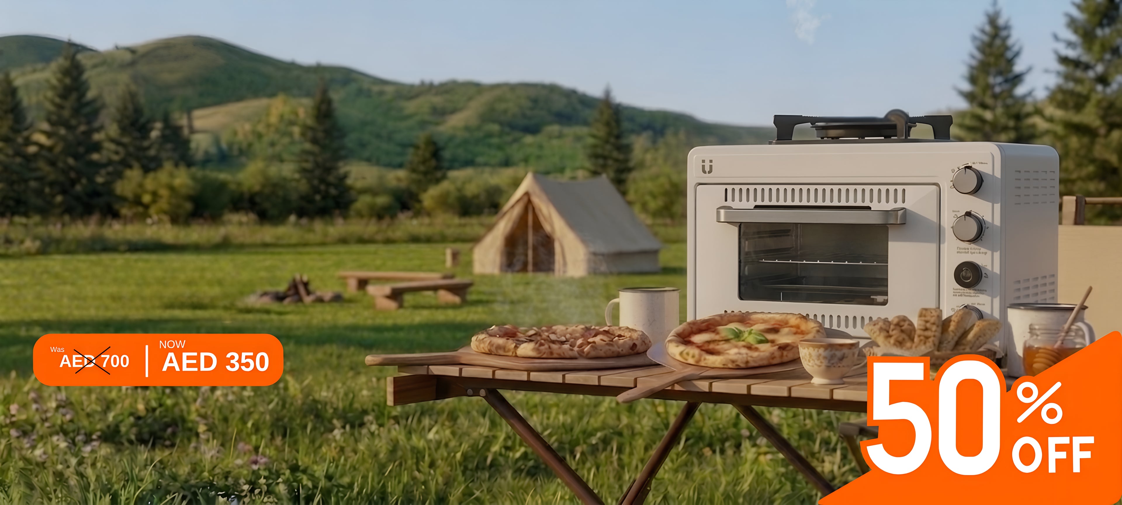 Picnic table with portable oven and stove, pizza, and drinks in a scenic outdoor setting with mountains and trees.